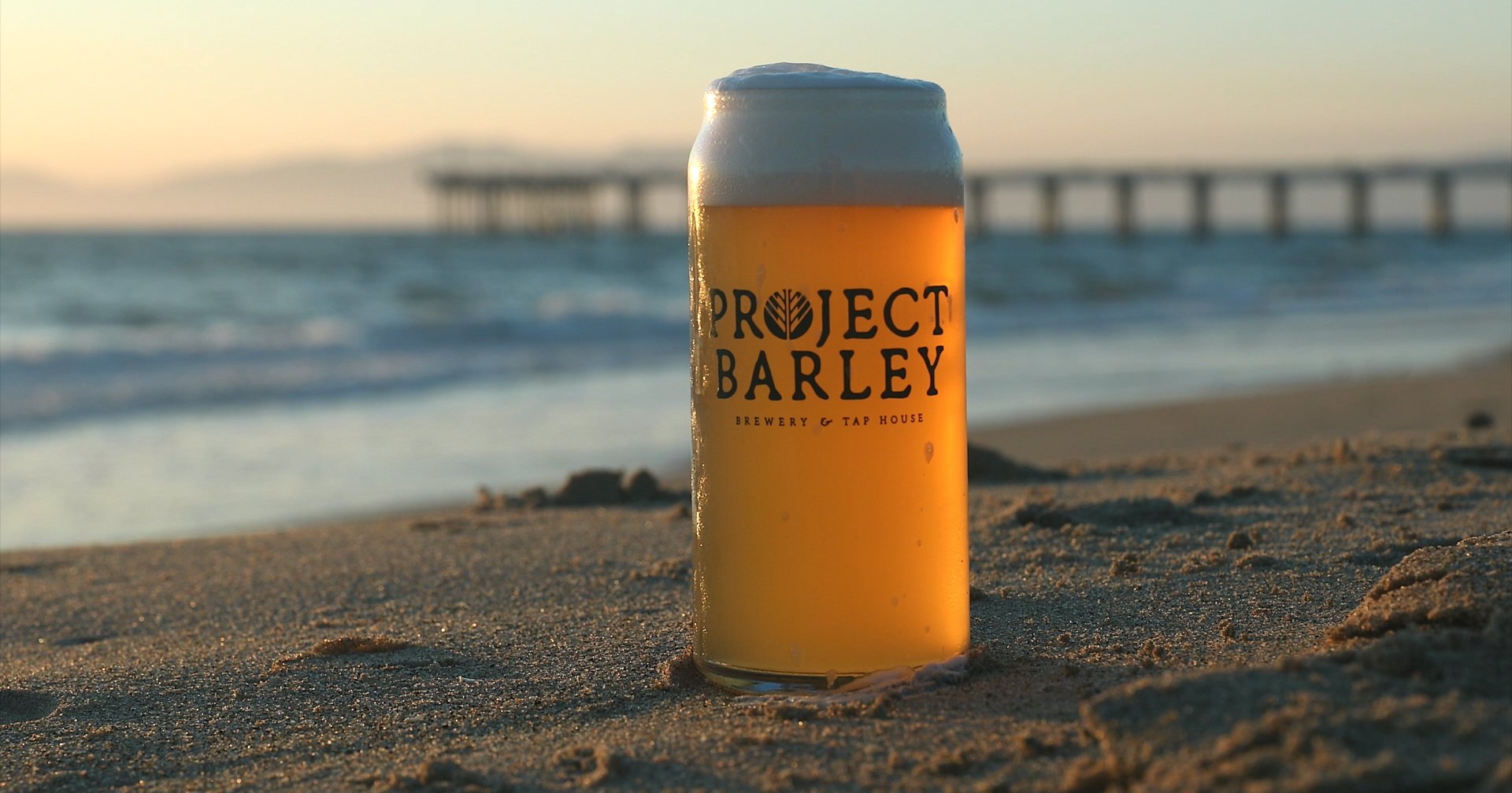 A Project Barley craft pour in the sand at sunset, with the pier behind